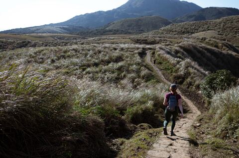 A tiny person with a backpack walking up a narrow dirt path that winds through dry grass, with a mountain and hills in the distance.